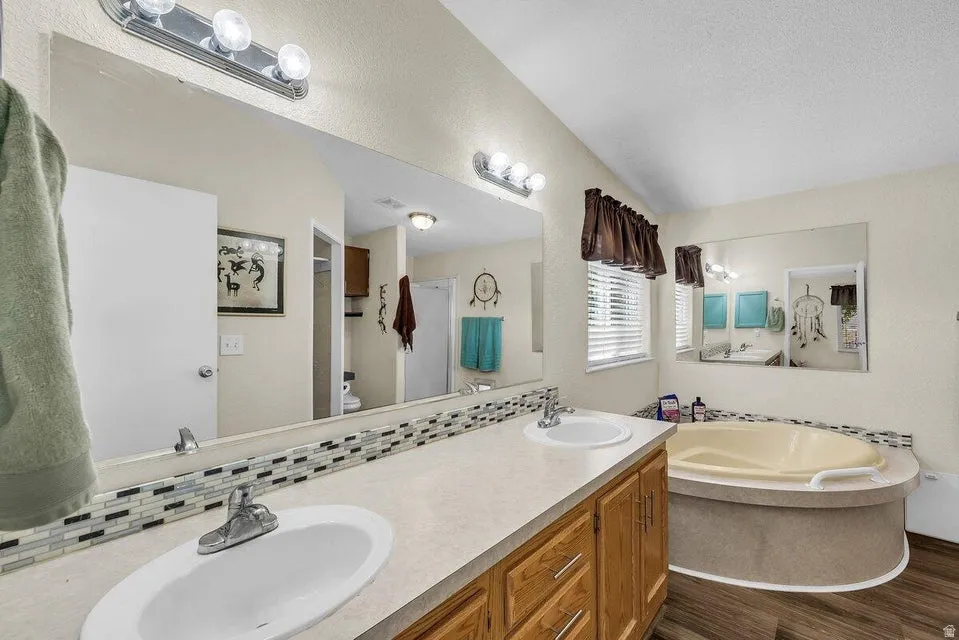 Bathroom featuring a dual vanity with a laminate countertop and white sinks, a large soaking tub, wood-finish flooring, and a window with blinds