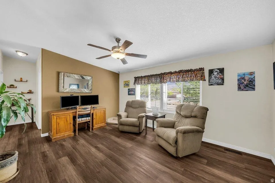 Living area featuring wood-finish flooring, a ceiling fan with integrated lighting, and multiple windows with blinds