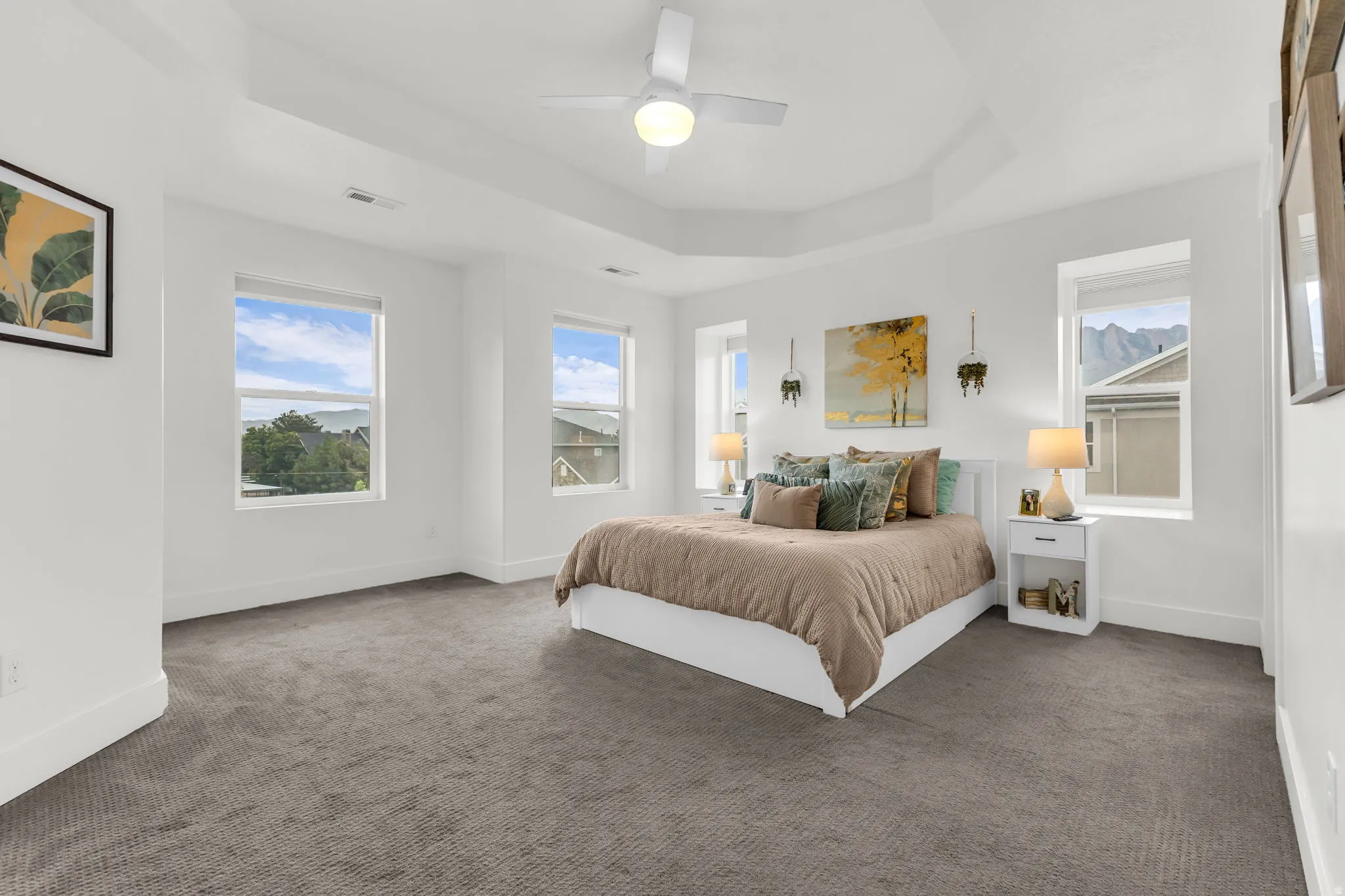 Spacious bedroom featuring a tray ceiling, ceiling fan, and multiple windows and plush carpet.