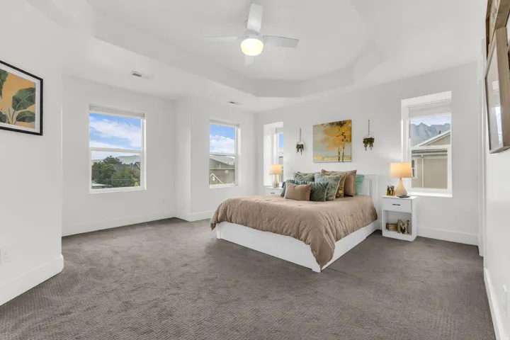 Spacious bedroom featuring a tray ceiling, ceiling fan, and multiple windows and plush carpet.