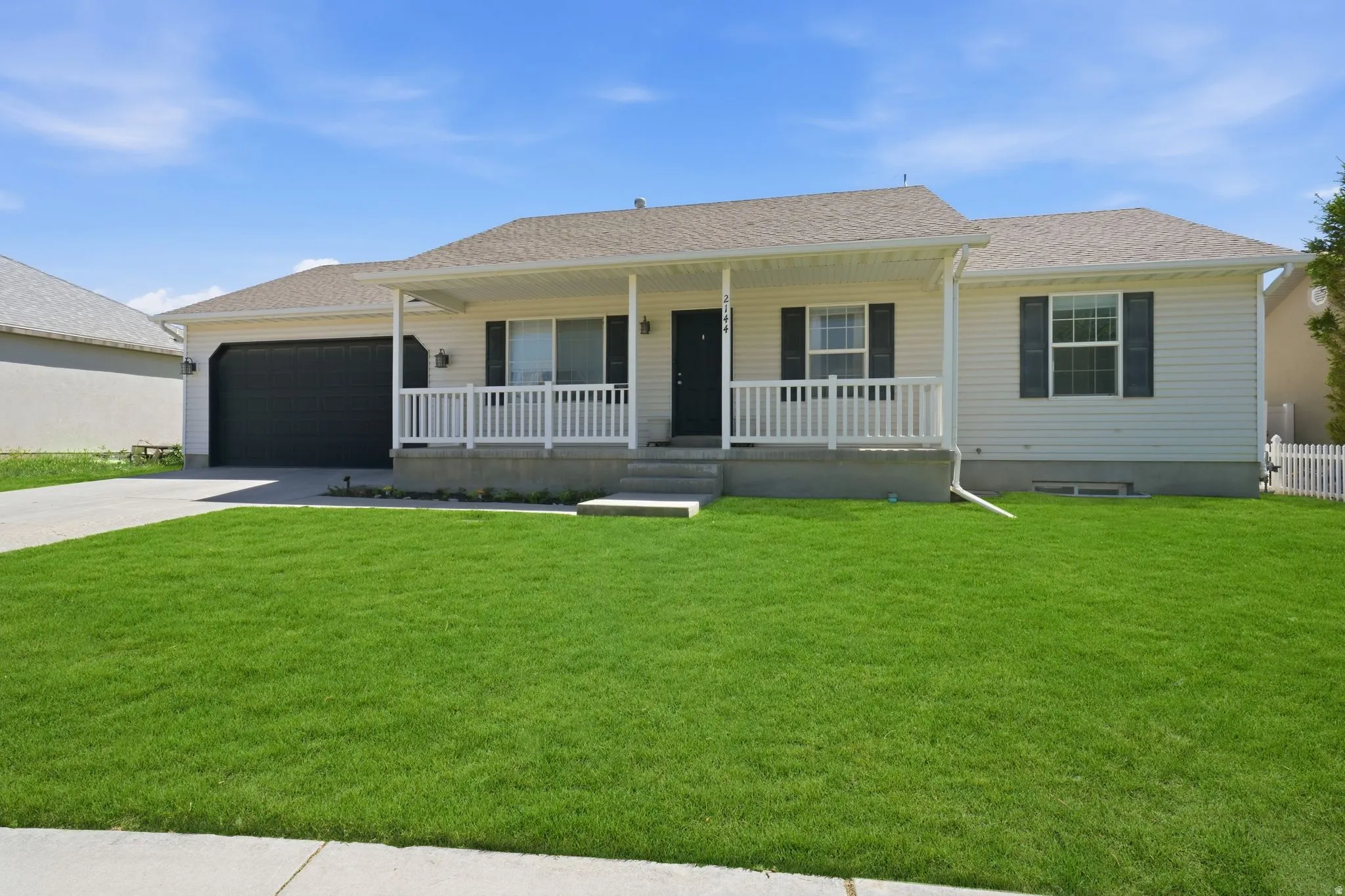 Ranch-style residence featuring light-colored siding, a covered front porch with white railings, a dark gray garage door, and a concrete driveway
