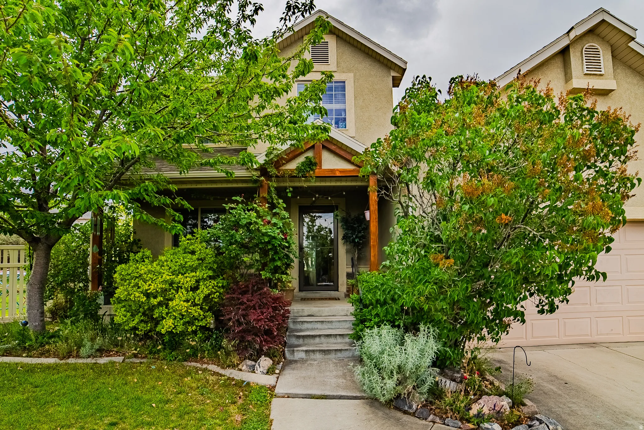 Stucco exterior featuring a covered front porch with exposed wood beams, a concrete pathway, mature trees, and established landscaping