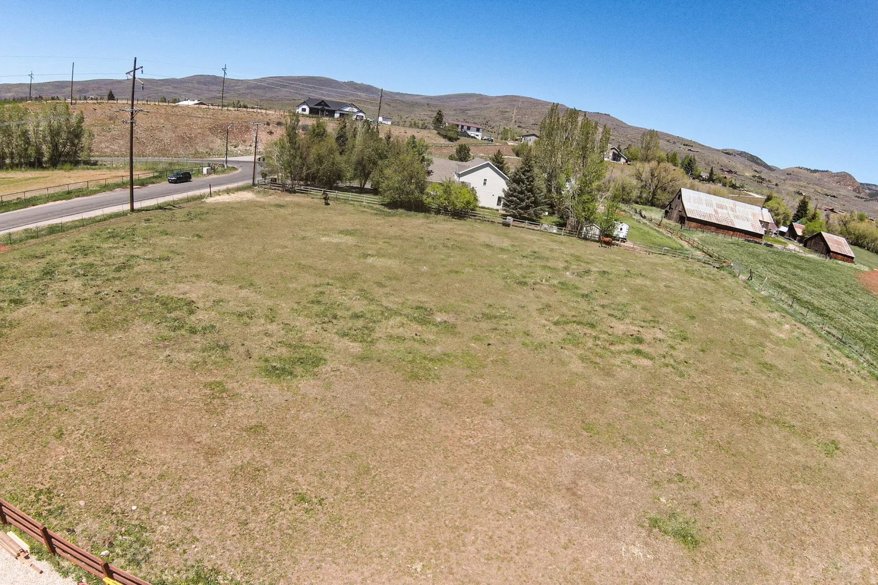 Expansive grassy land featuring a paved road, scattered trees, and distant residential structures