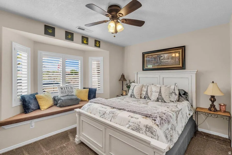 Bedroom featuring a bay window with a built-in cushioned window seat, white plantation shutters, a ceiling fan with integrated lighting, and neutral wall tones