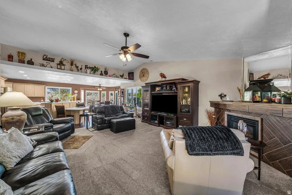 Spacious living area featuring vaulted ceilings, a ceiling fan, plush carpet flooring, a built-in fireplace with a wood-finish surround, and a broad pass-through counter to the kitchen