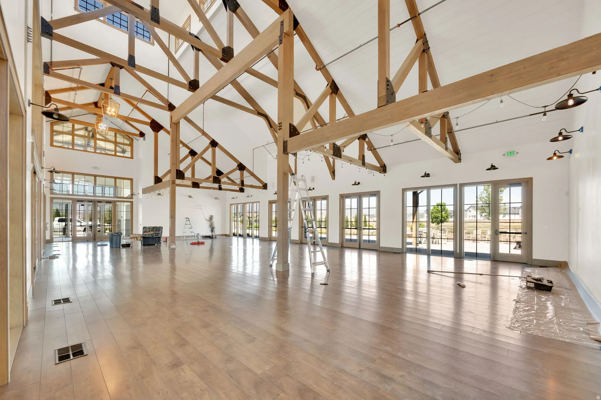 Spacious interior featuring exposed timber trusses, high vaulted ceilings, expansive wood-finish flooring, multiple sets of French doors, and decorative wall-mounted lighting fixtures