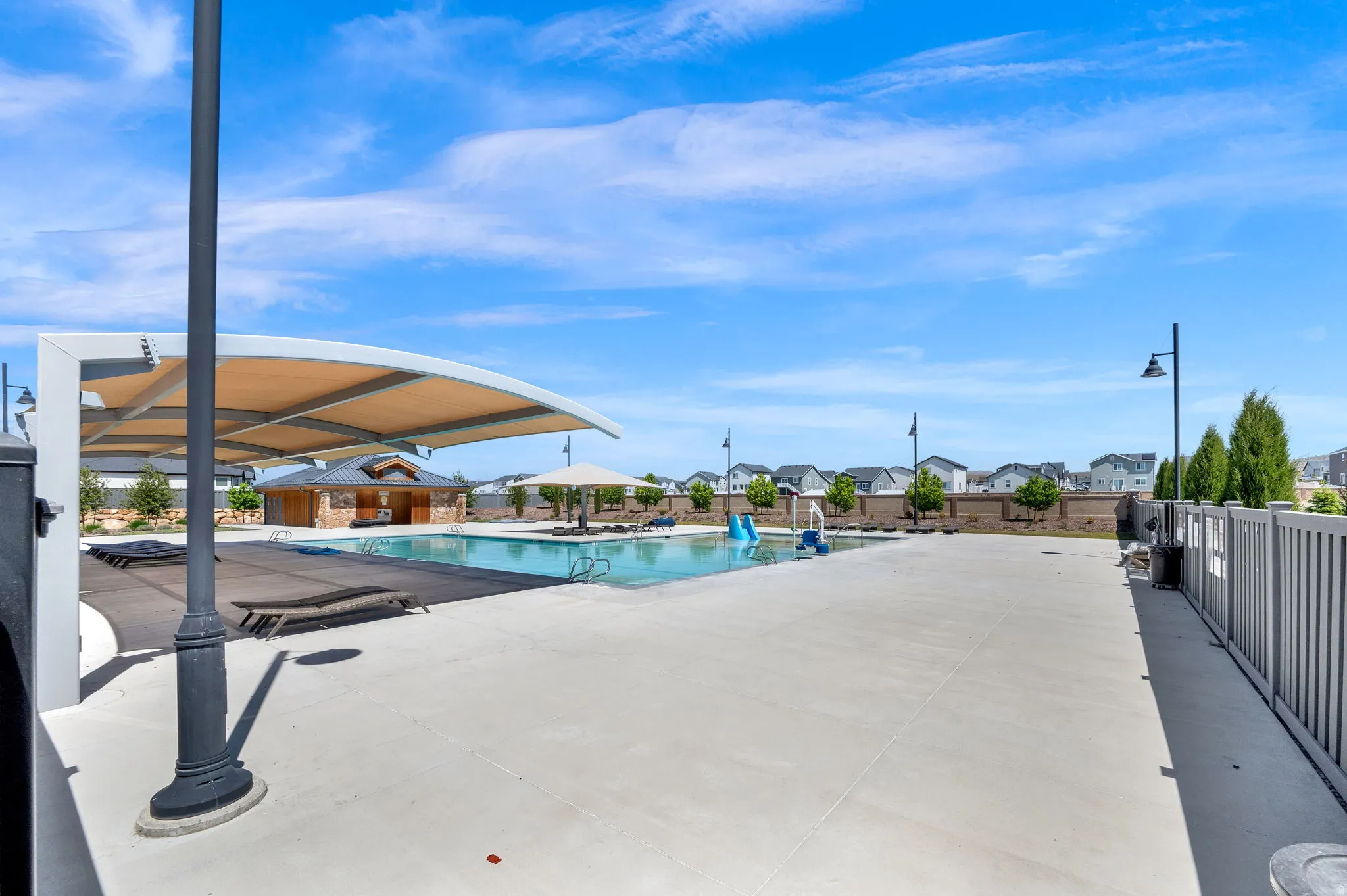 Community swimming pool featuring a curved shade structure, a separate pool house building with stone detailing, an adjacent splash pad with blue and white water features, and expansive concrete decking