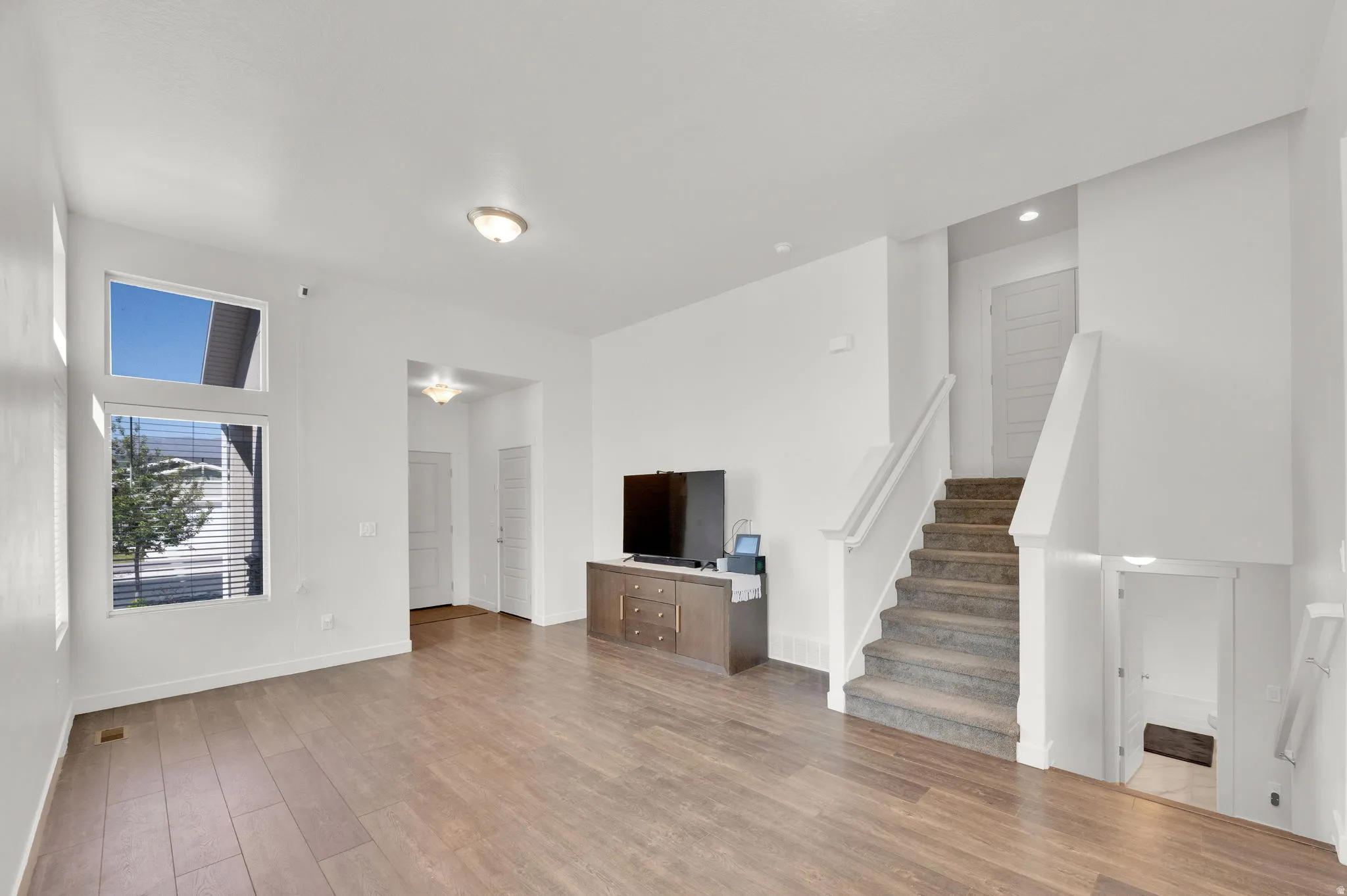 Spacious living area featuring wood-finish flooring, high ceilings, multiple windows, and a carpeted staircase with a white banister