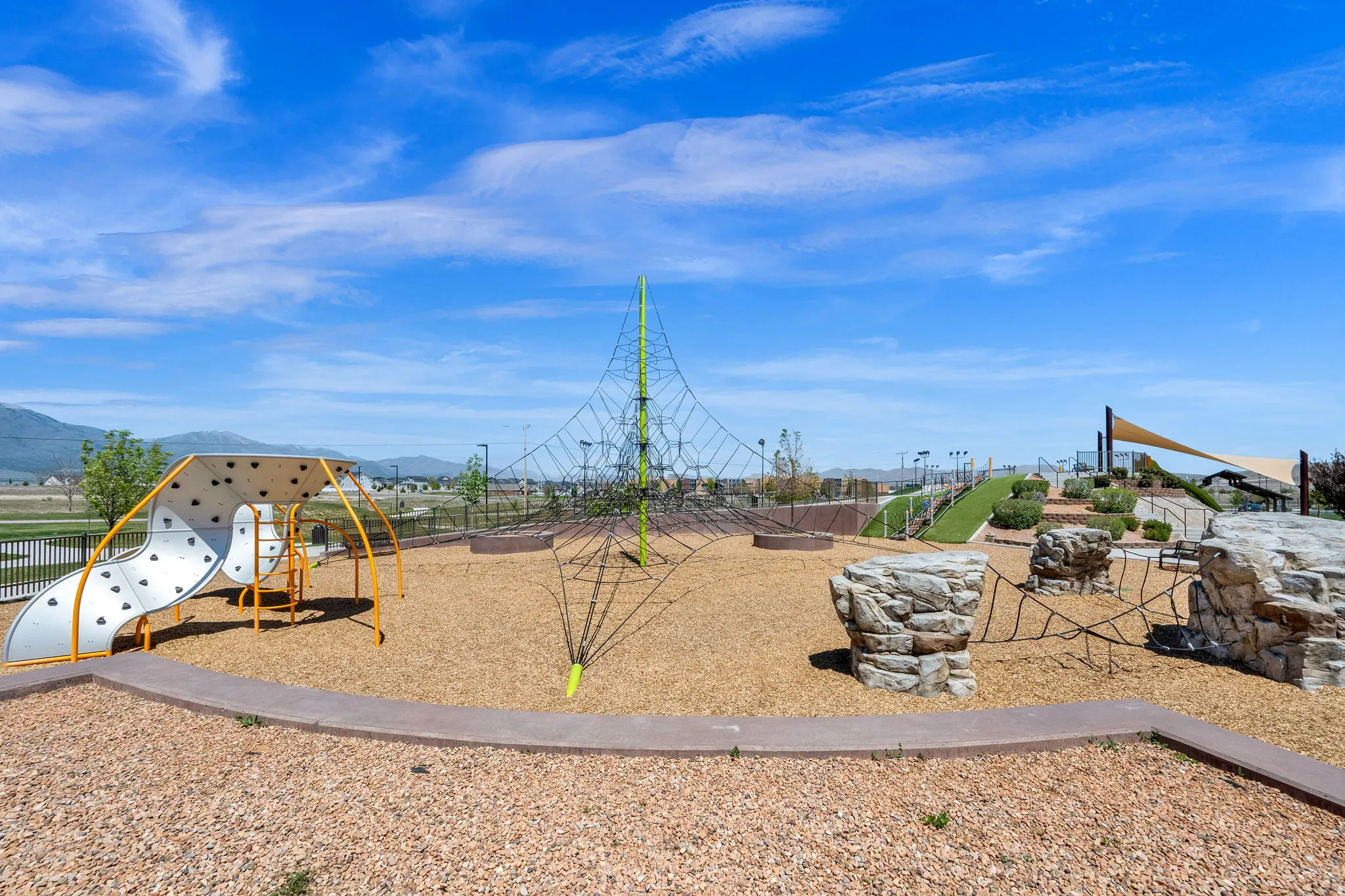 Community playground featuring varied play structures, a large climbing net, rock-like climbing elements, a slide, and a shade sail