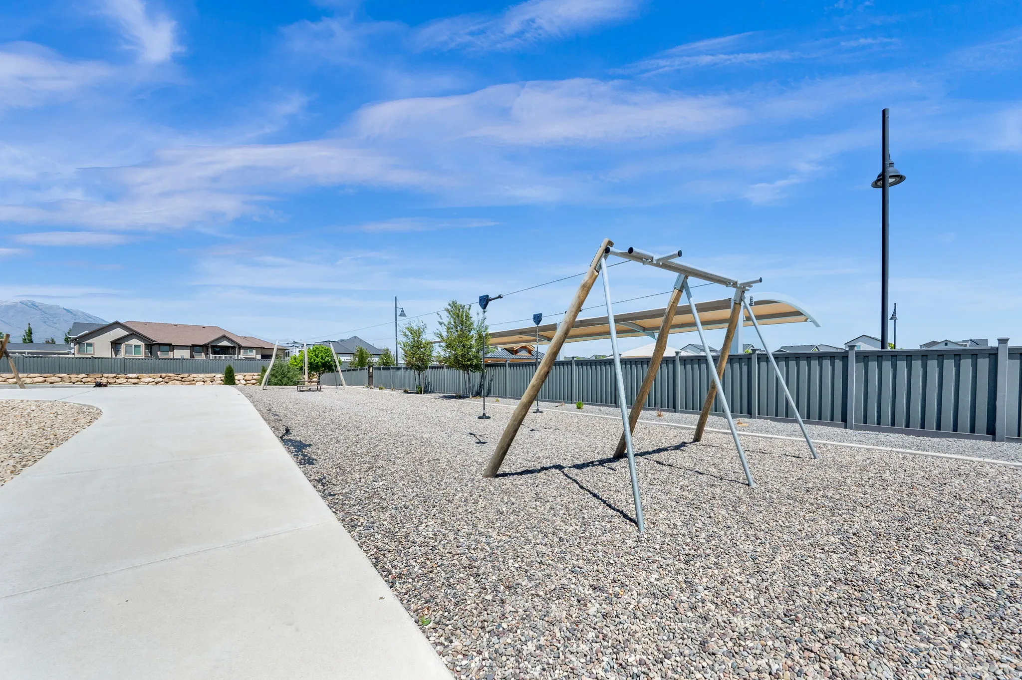Outdoor recreational area featuring a gravel surface, playground equipment with sun shades, a concrete pathway, a privacy fence, and distant mountain scenery