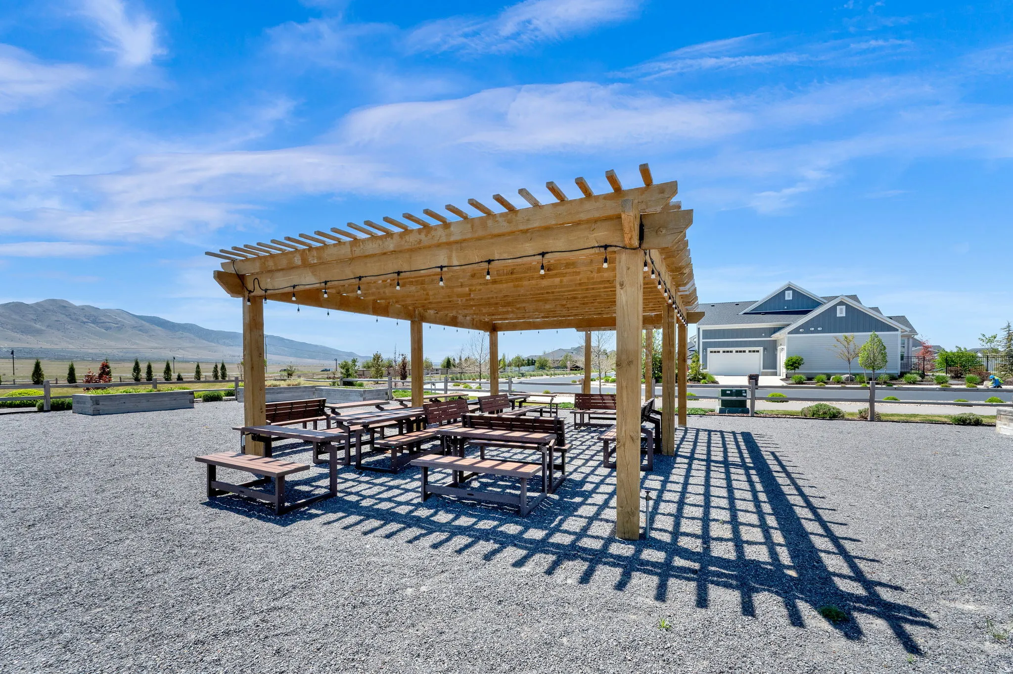 Expansive outdoor pergola structure with string lighting, multiple picnic tables, and benches, set on a gravel surface