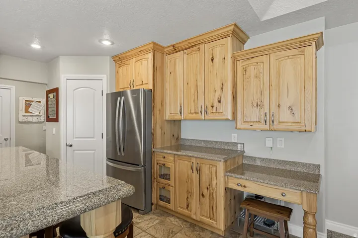 Kitchen featuring natural wood cabinetry, light-toned granite countertops, a stainless steel refrigerator, recessed lighting, and tile flooring