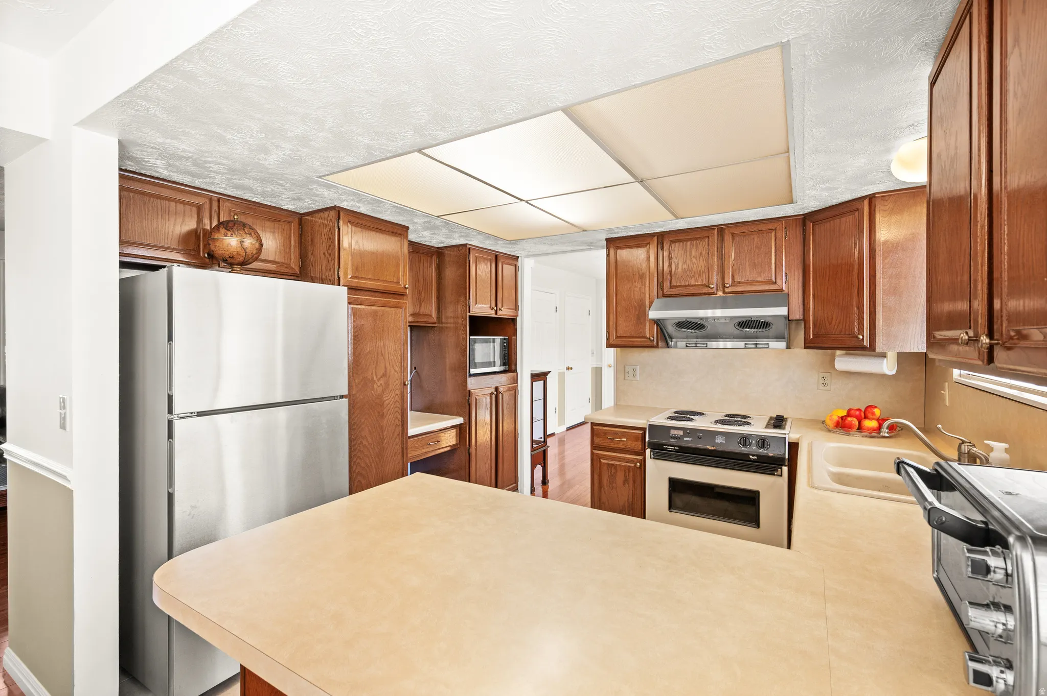 Kitchen featuring wood cabinetry, light-toned countertops, a built-in electric range, a stainless steel refrigerator, and a recessed ceiling light fixture