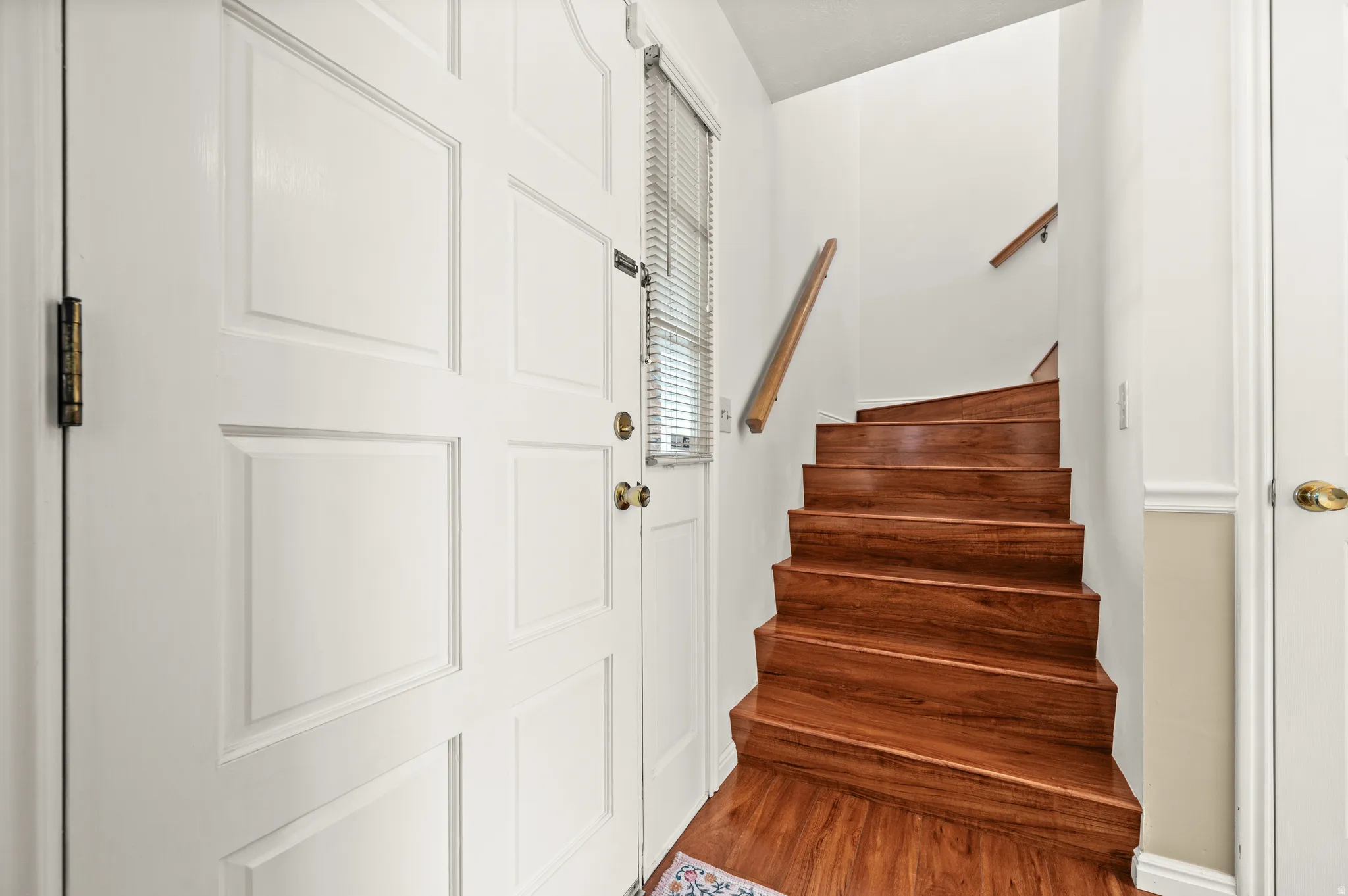 Wood-finish staircase with a natural wood handrail, white paneled walls, and a white paneled door with brass hardware