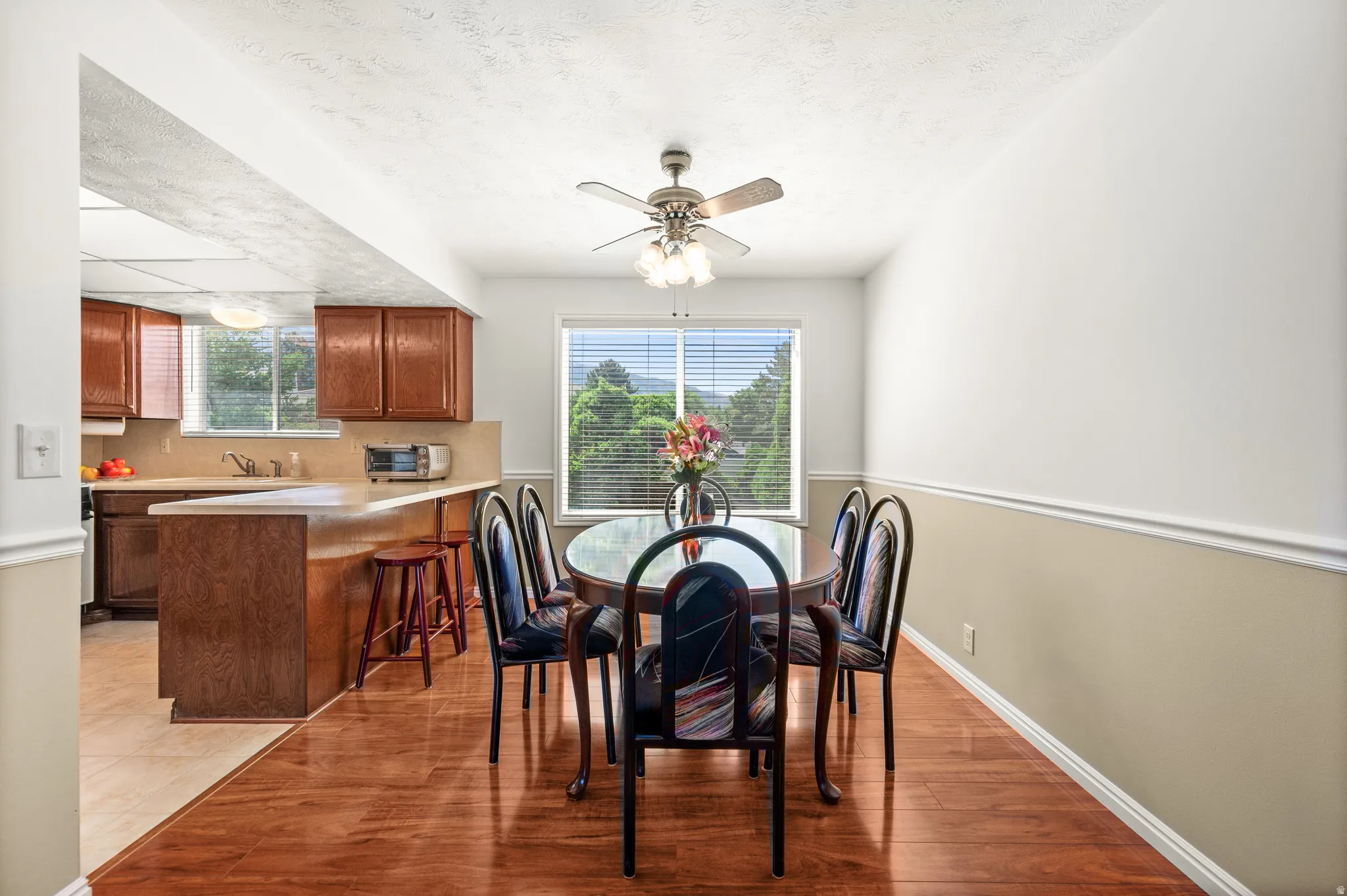 Dining area featuring wood-finish flooring, a ceiling fan with light fixture, and a window with blinds overlooking greenery