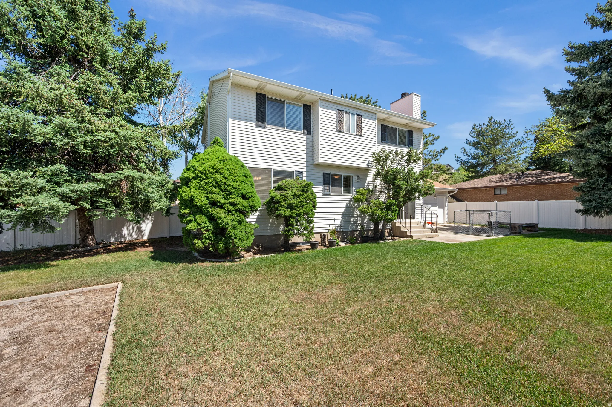 Expansive grassy yard area featuring mature evergreen trees, a concrete patio, and white privacy fencing