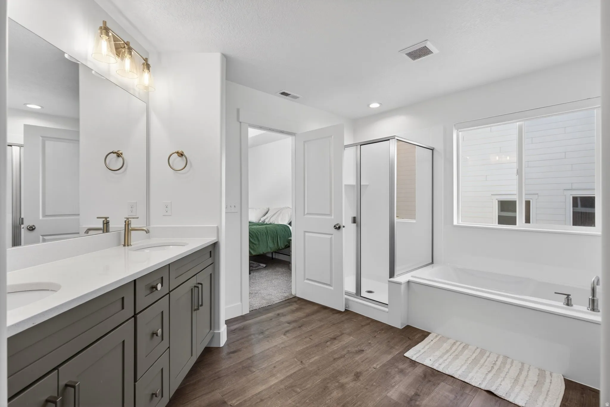 Spacious primary bathroom featuring a dual vanity with a white countertop, wood-finish flooring, a separate walk-in shower with glass enclosure, a soaking tub, and a large window