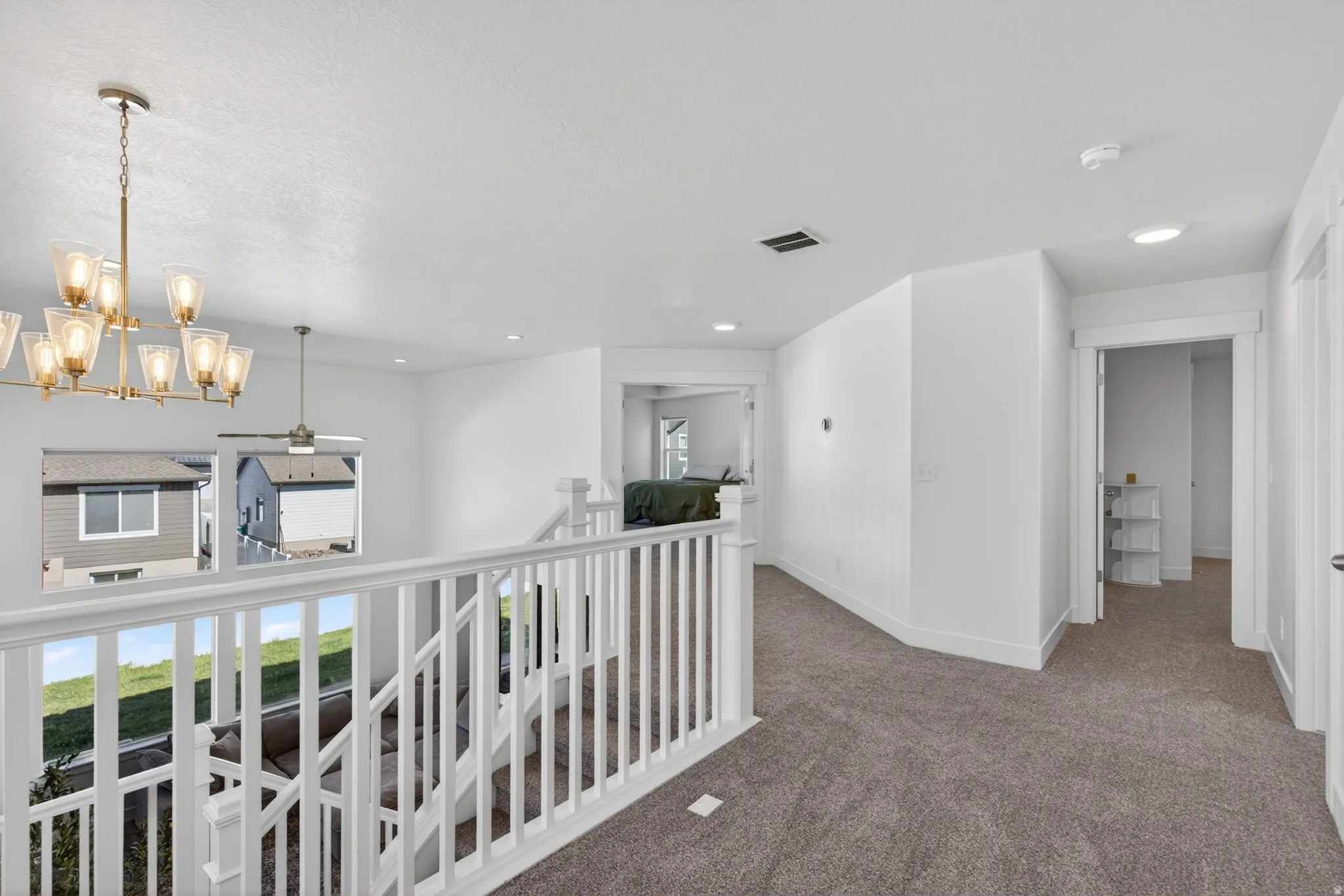 Carpeted hallway with white banister overlooking a lower level, featuring recessed lighting, a modern chandelier, and a ceiling fan