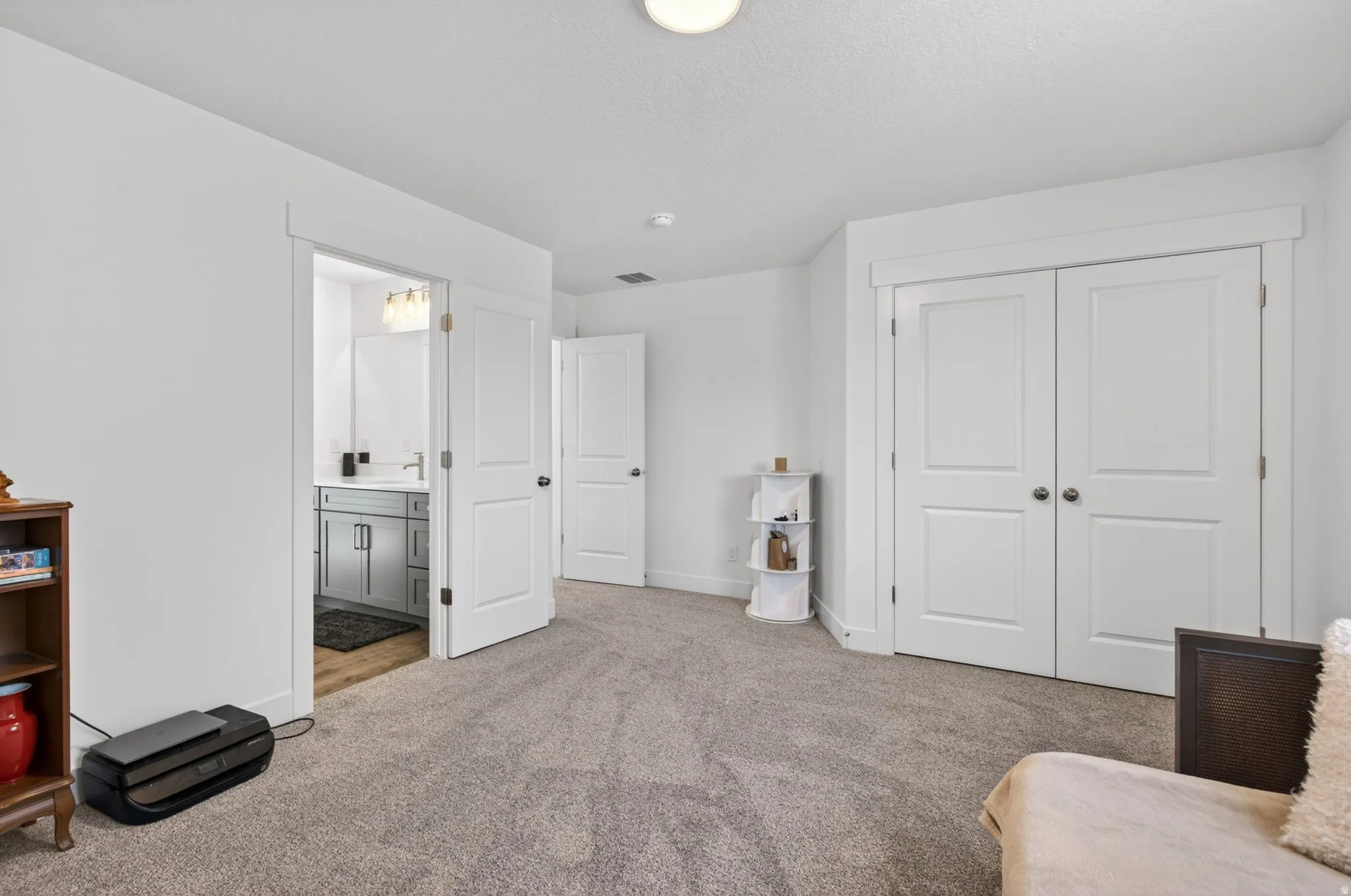Carpeted room with white walls, featuring a double-door closet with white paneled doors, and an entry to a bathroom with a gray vanity