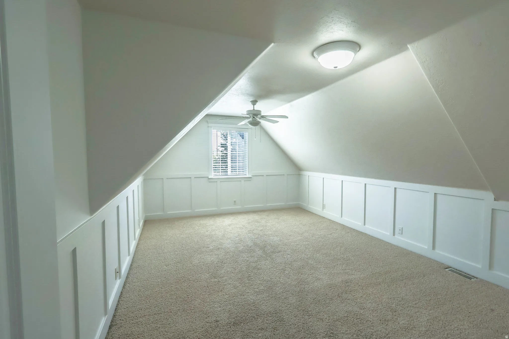 Room with sloped ceilings featuring wall paneling, neutral carpeting, and a window with blinds