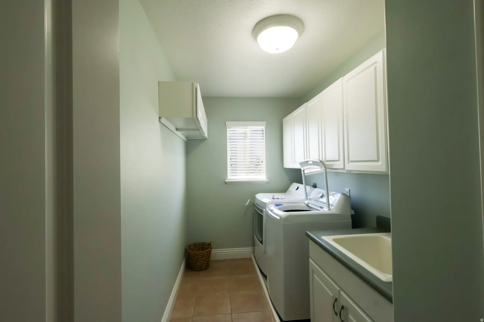 Dedicated laundry room featuring tile flooring, white shaker-style cabinetry, a built-in utility sink with laminate countertop, a single window with blinds, and flush-mount ceiling lighting