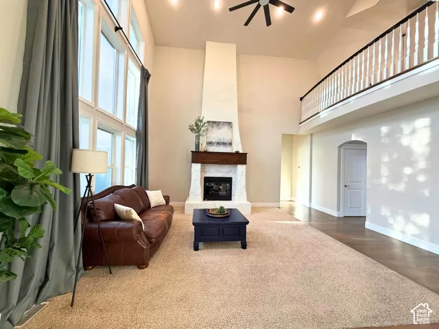 Soaring ceilings with a decorative ceiling fan, a fireplace with a wood-mantel and stone surround, a prominent floor-to-ceiling window arrangement, a lofted area with a white spindle railing, and a blend of carpet and hard-surface flooring