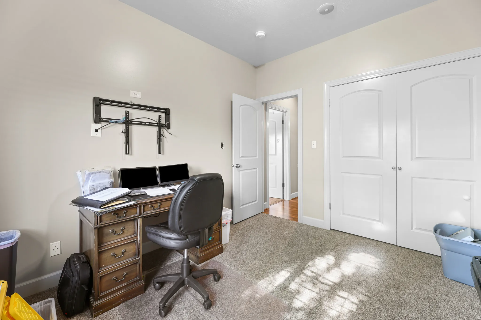 Carpeted room featuring a wall-mounted bracket, a pair of white bifold doors with arched paneling, and an interior door leading to a hallway with wood-finish flooring