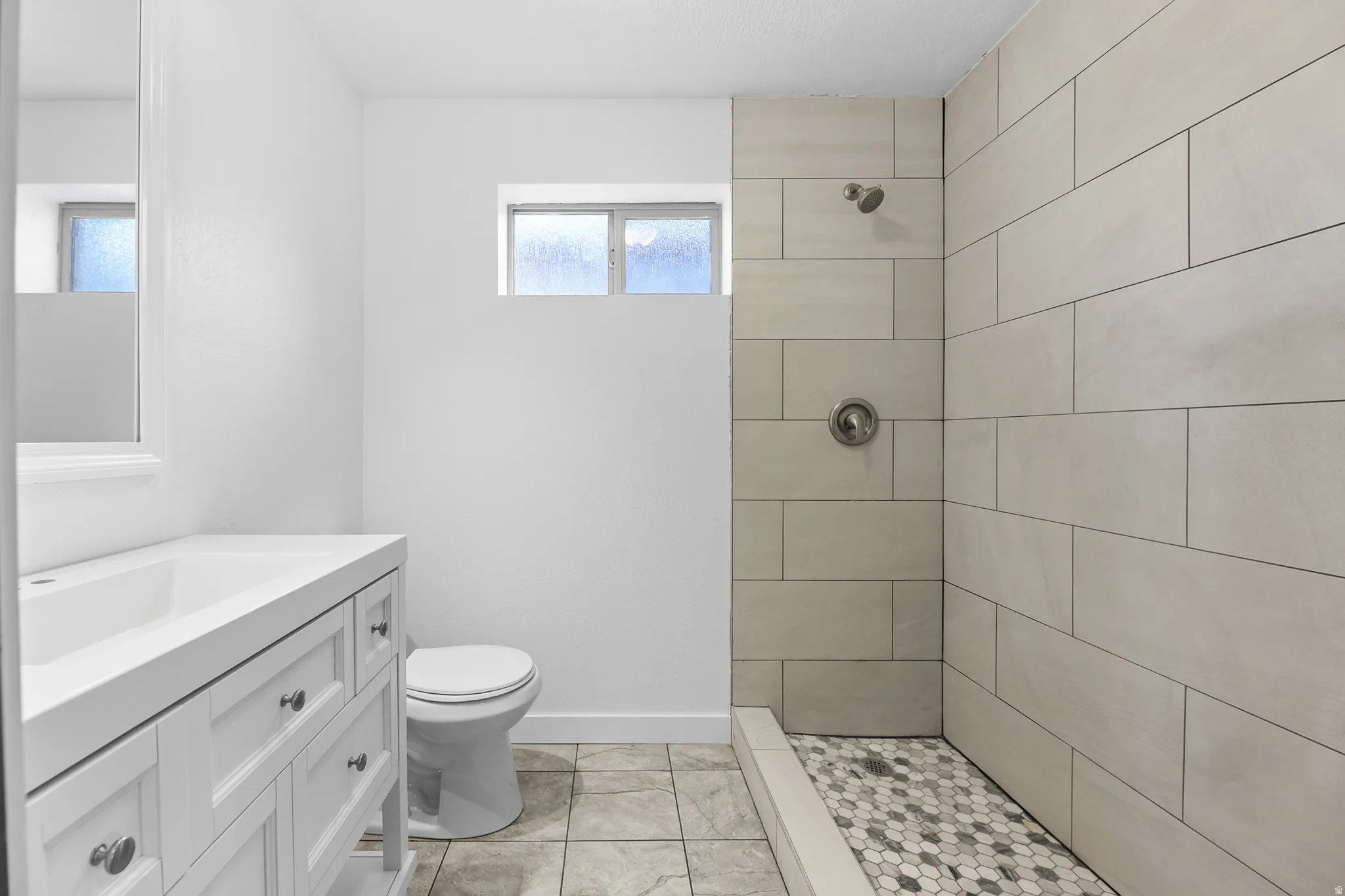 Shower with rectangular wall tiles and a hexagonal mosaic floor, white vanity with integrated sink, and a high-set window