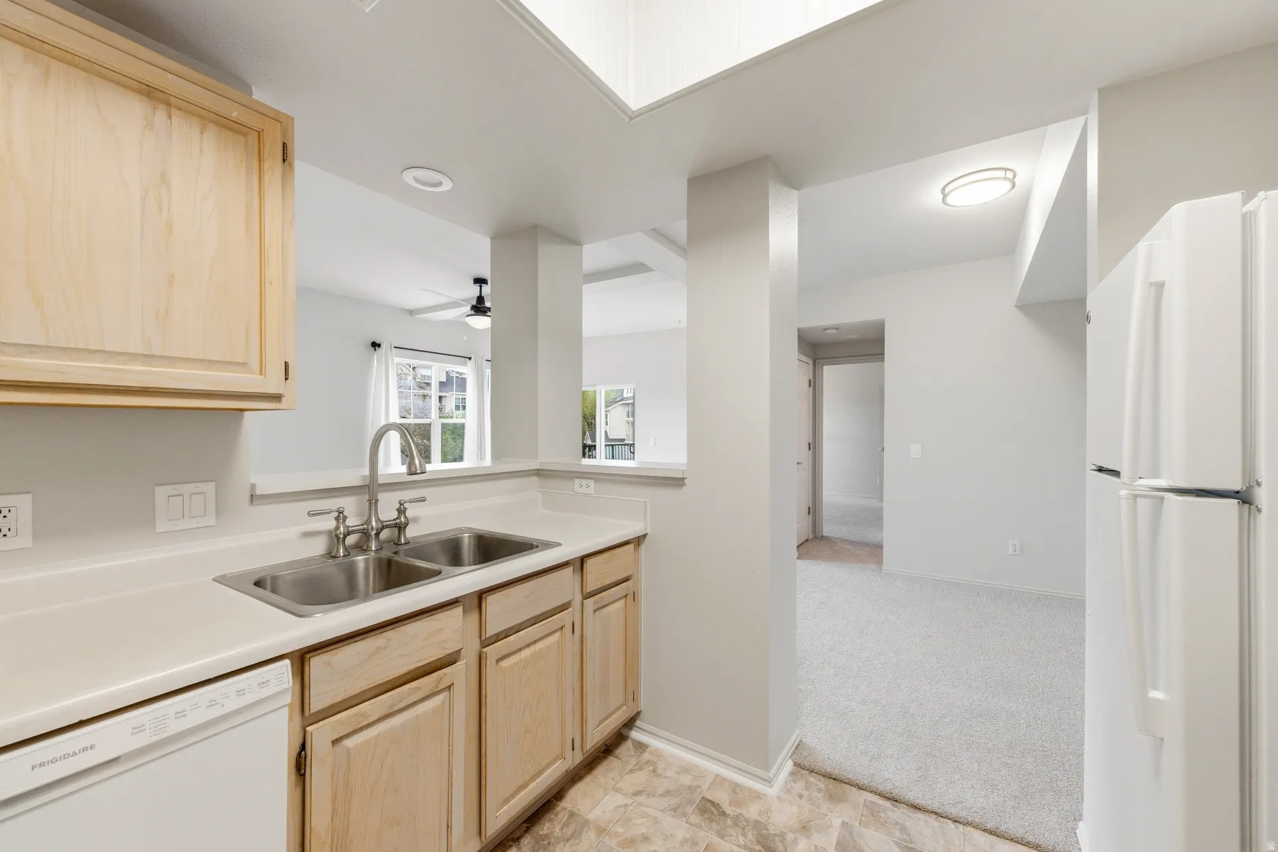 Kitchen featuring light wood-finish cabinetry, solid surface countertops, a stainless steel double basin sink with a chrome faucet, and integrated white appliances