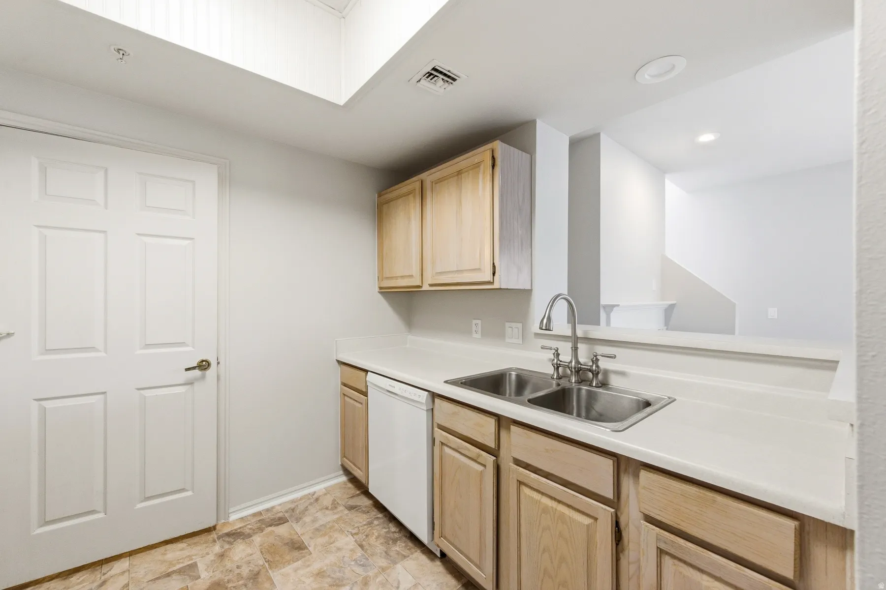 Kitchen area featuring light wood-finish cabinetry, a double basin stainless steel sink with a gooseneck faucet, a white dishwasher, light-toned countertops, and patterned floor tiling