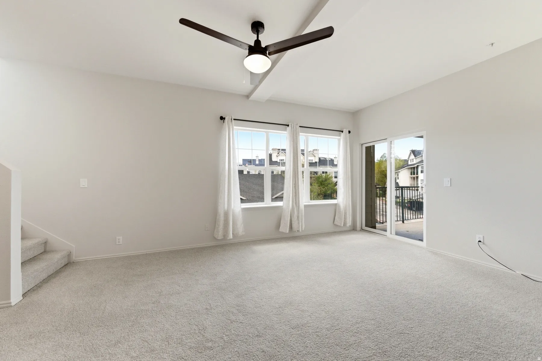Spacious carpeted room featuring a ceiling fan with integrated lighting, multiple windows, and a sliding glass door opening to a balcony