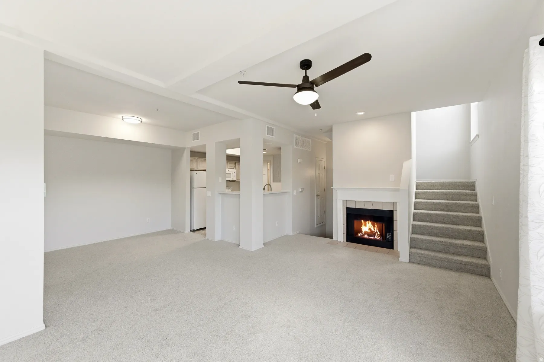 Carpeted living area featuring a fireplace with a white mantel, ceiling fan, and a light-toned kitchen with white appliances