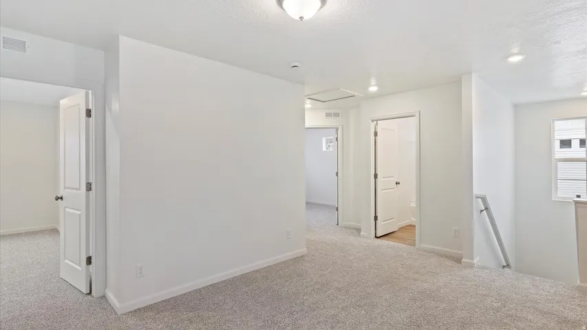 Carpeted hallway with white walls and trim, featuring multiple paneled doors, ceiling-mounted lighting fixtures, and a handrail leading to a lower level