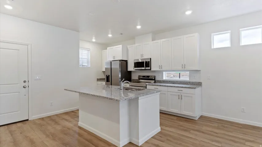 Kitchen featuring a central island with a sink, stainless steel appliances, white cabinetry, speckled countertops, and wood-finish flooring