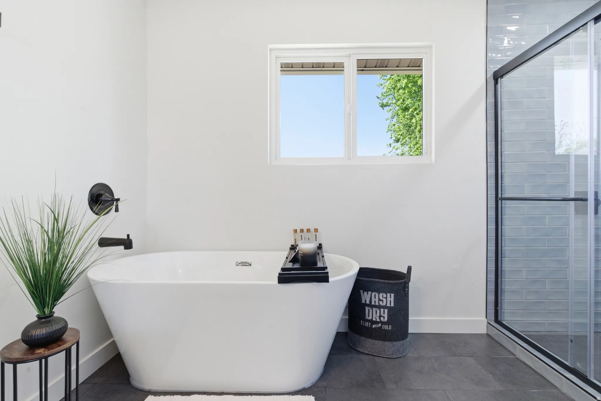 Freestanding soaking tub with matte black fixtures, framed by a sliding window and a glass-enclosed shower featuring subway tile