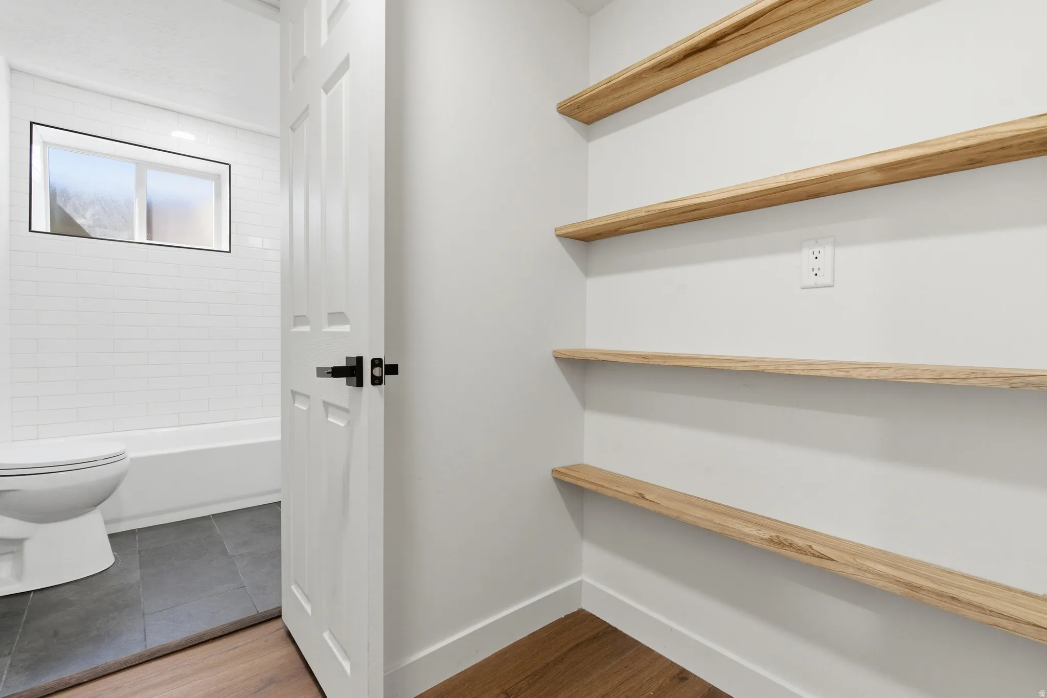 White interior featuring four wood-finish floating shelves, a white paneled door with black hardware, and wood-finish flooring