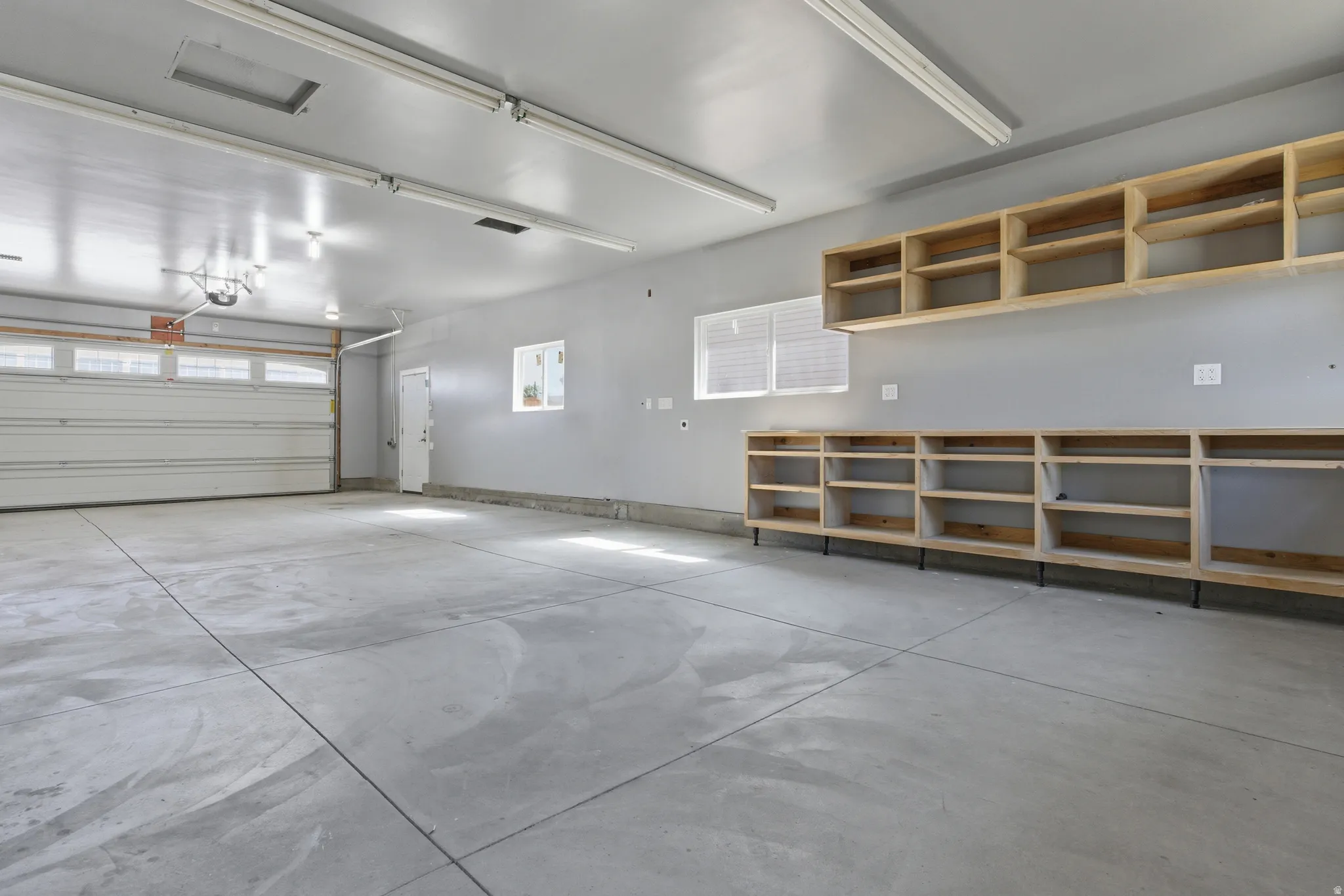 Spacious garage featuring a concrete floor, built-in wooden shelving, multiple windows, overhead fluorescent lighting, and a roll-up garage door