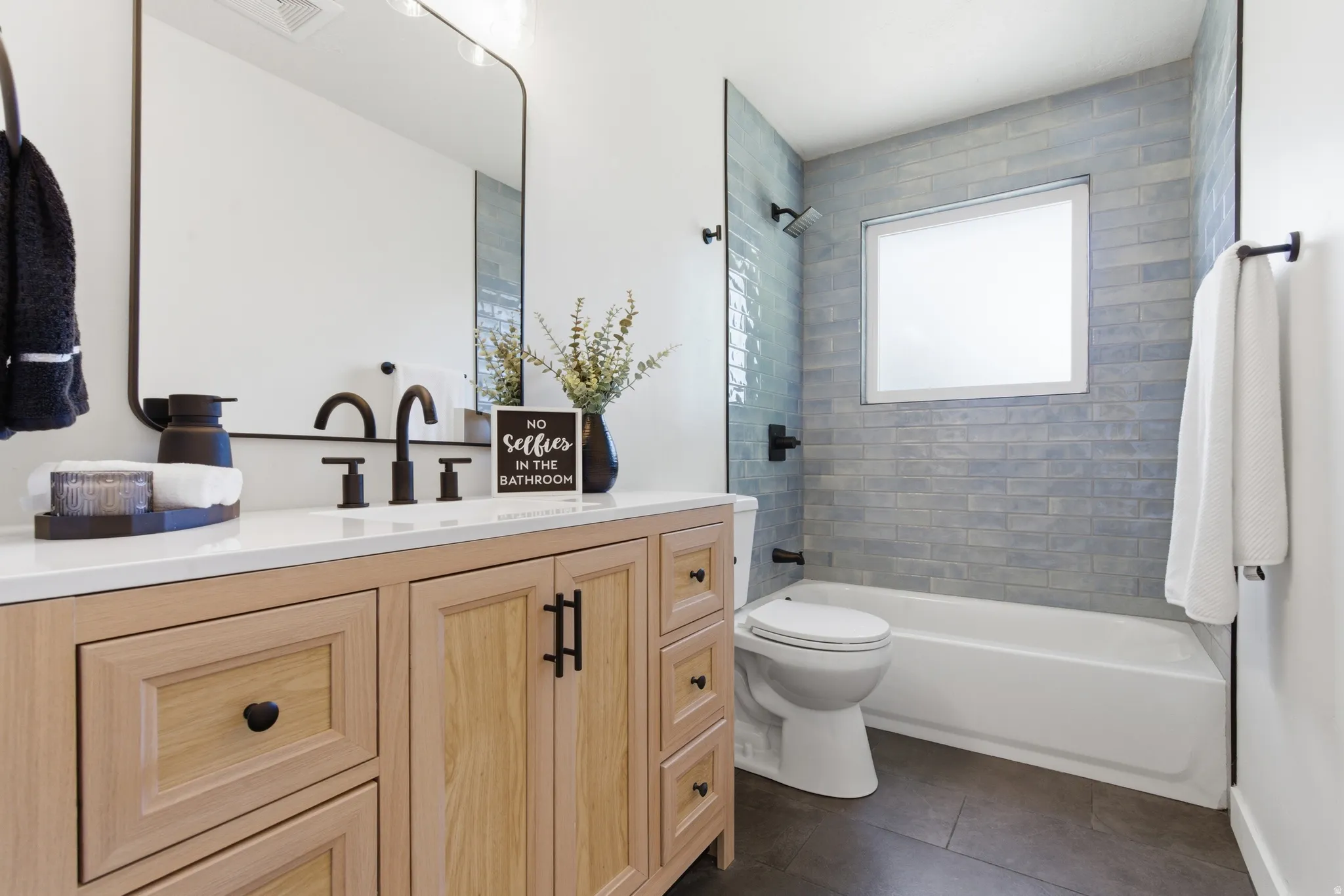 Bathroom featuring a wood-finish vanity with a white countertop, matte black hardware and fixtures, a rectangular mirror, a subway tile shower surround, and dark floor tiles