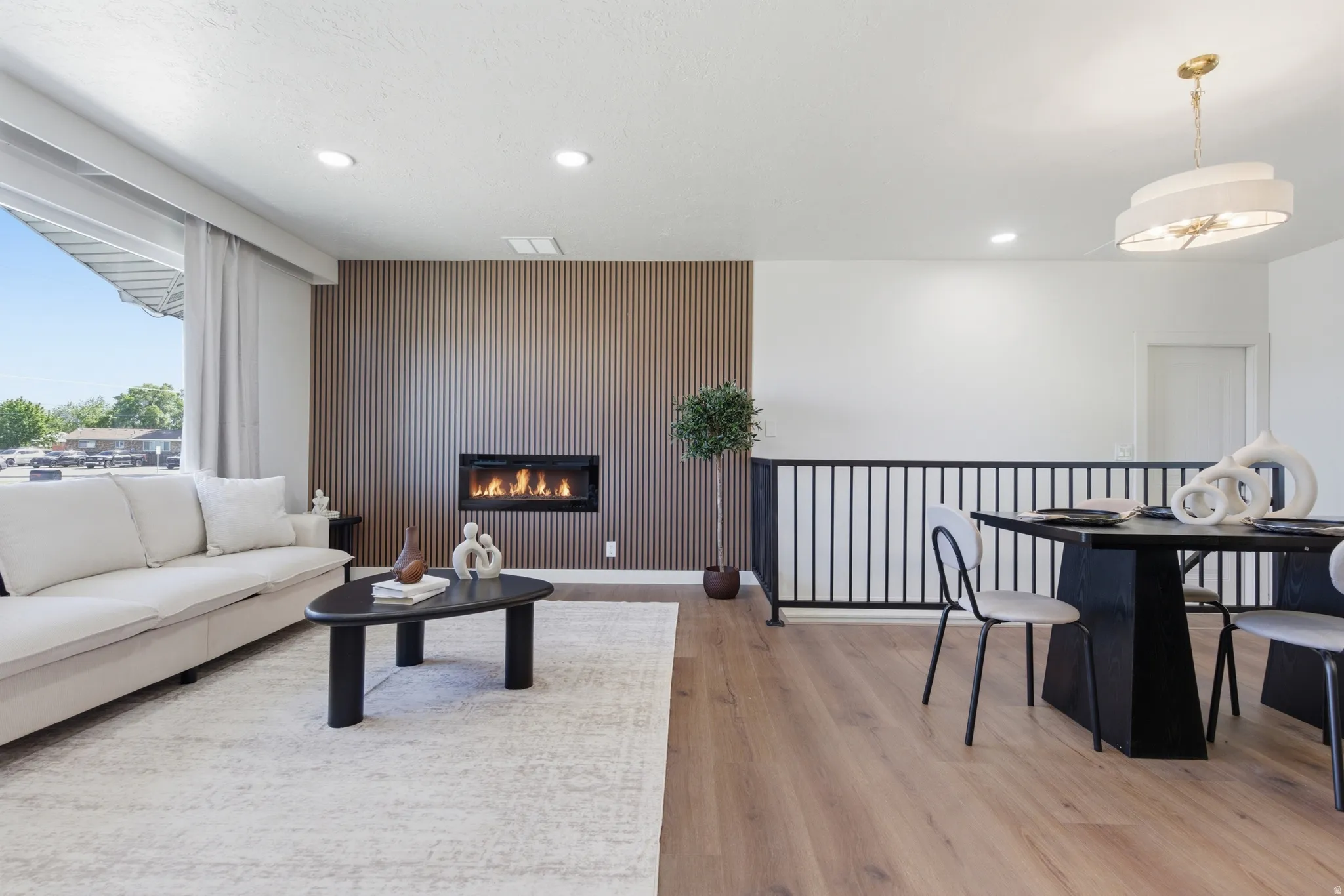 Open-concept living space featuring wood-finish flooring throughout, a modern linear fireplace with a slatted wood accent wall, recessed lighting, and a contemporary dining chandelier