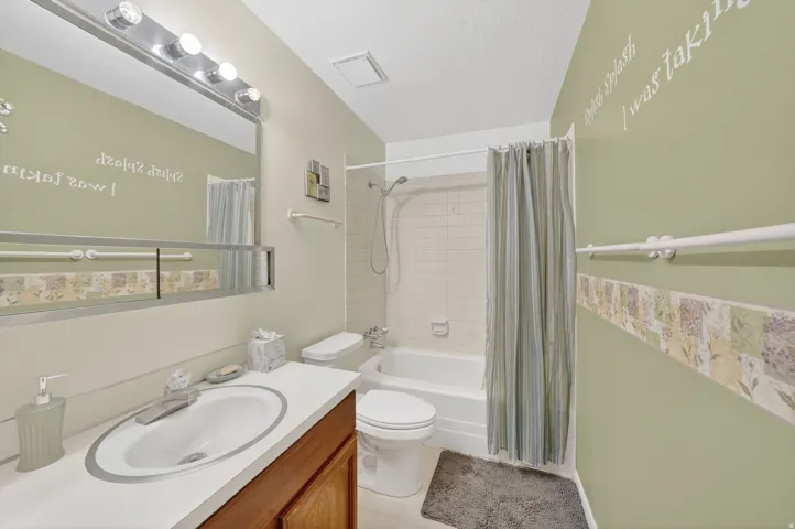 Full bathroom featuring a white vanity with an integrated sink and wood-finish cabinetry, a large vanity mirror with overhead lighting, a white toilet, and a shower-tub combination with white tile surround