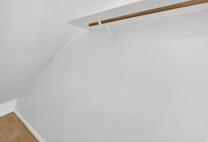 Sloped wall closet featuring a wooden hanging rod, white shelf with supports, white baseboards, and tan carpeting