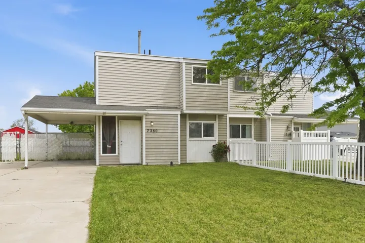 Exterior featuring a carport, front lawn, white picket fence, and a neutral-toned facade with multiple windows