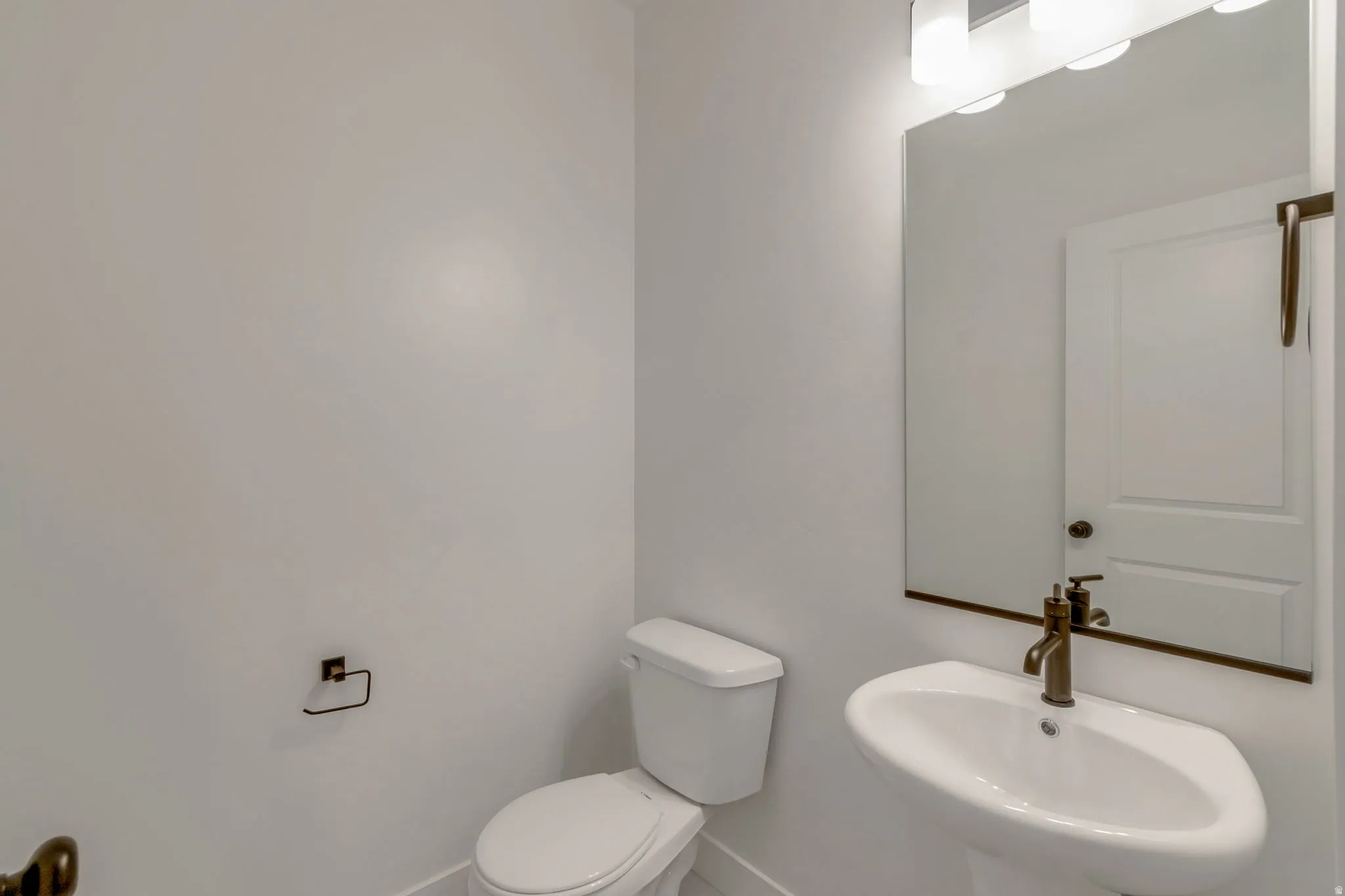 Pedestal sink with bronze-finish faucet, mirror with overhead vanity lighting, white toilet, and bronze-finish toilet paper holder