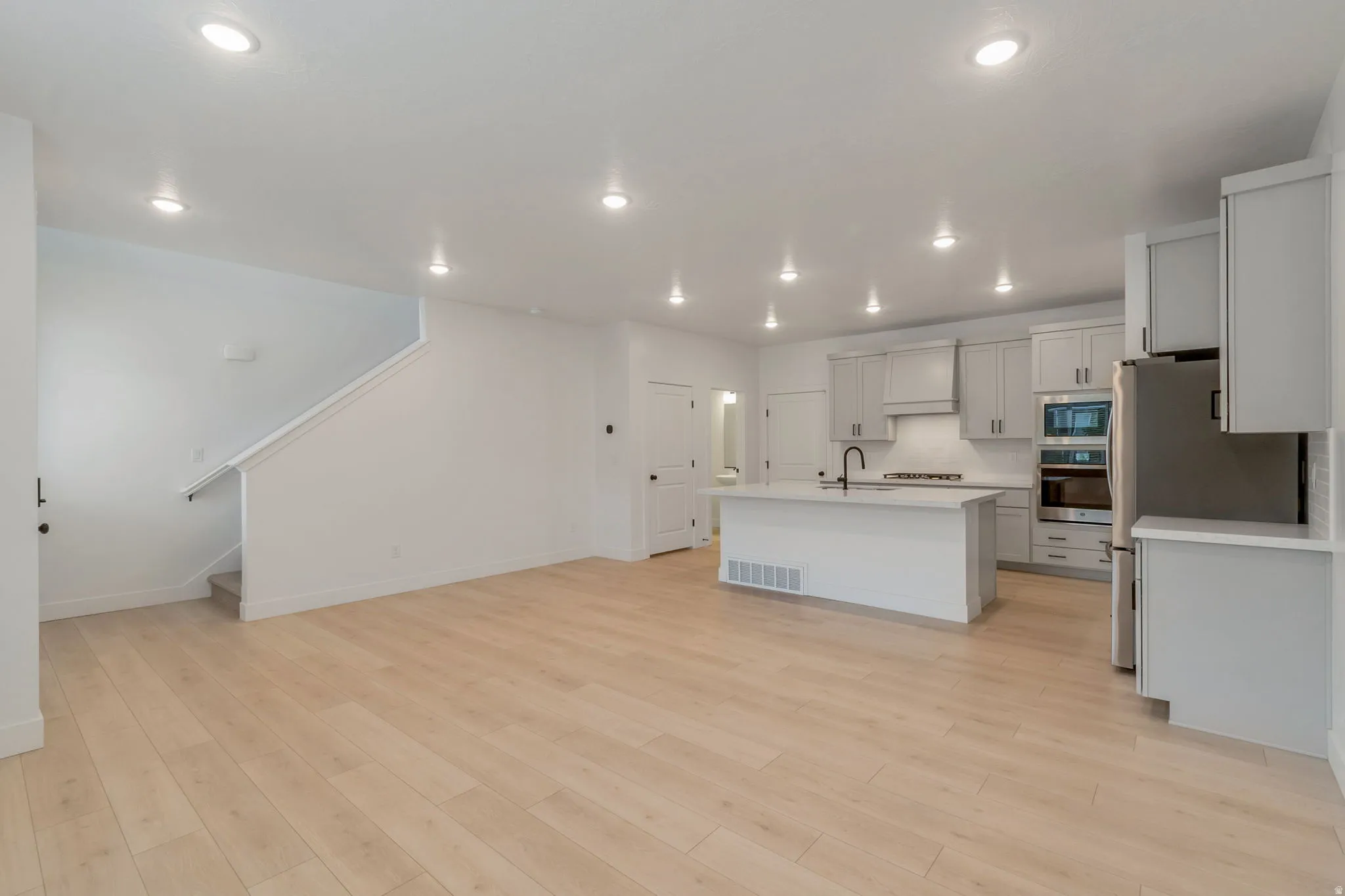 Open-concept living space featuring wood-finish flooring, a modern kitchen island with an integrated sink and matte black faucet, light gray cabinetry, stainless steel appliances, and recessed lighting throughout