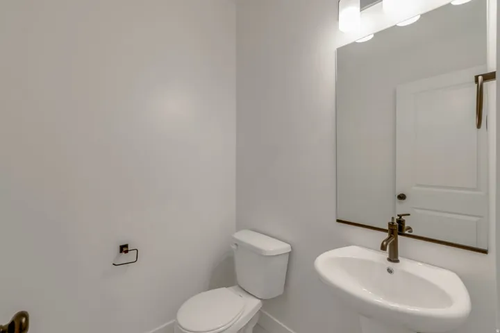 Pedestal sink with bronze-finish faucet, mirror with overhead vanity lighting, white toilet, and bronze-finish toilet paper holder