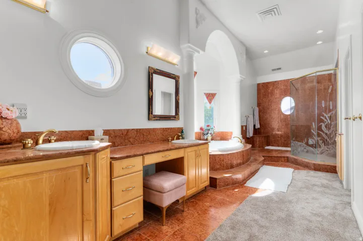Expansive bathroom featuring a dual vanity with a makeup station, a circular window, a tiled soaking tub, and a glass-enclosed shower with custom etched details