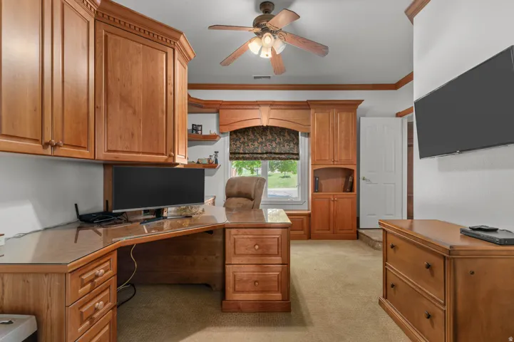 Dedicated office space featuring extensive built-in wood cabinetry and shelving, a custom L-shaped wood desk with a glass top, and a ceiling fan with integrated lighting