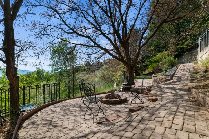Paver patio featuring a built-in fire pit, surrounded by mature trees and a black metal fence