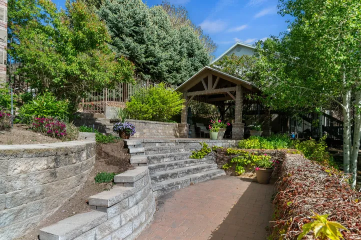 Terraced outdoor space featuring a stone paver patio, block retaining walls, and a timber-framed gazebo with stone pillars