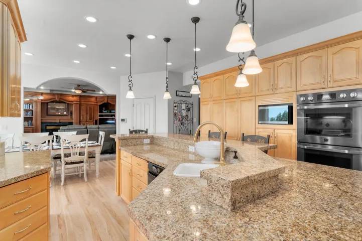 Kitchen featuring a large island with an undermount sink and golden-toned faucet, light wood cabinetry, recessed lighting, pendant lights, and stainless steel double ovens